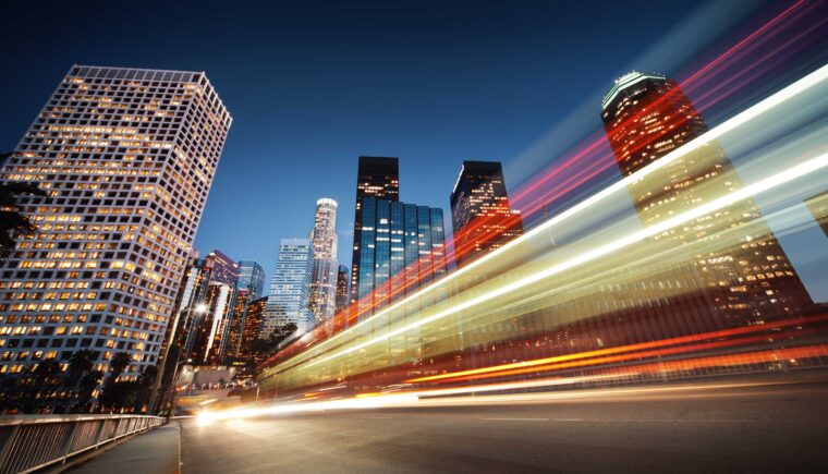 A nighttime cityscape with illuminated skyscrapers in downtown Los Angeles, featuring light trails from moving vehicles captured using long exposure, creating dynamic streaks of red, white, and yellow across the foreground.