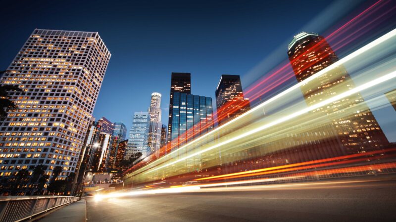 A nighttime cityscape with illuminated skyscrapers in downtown Los Angeles, featuring light trails from moving vehicles captured using long exposure, creating dynamic streaks of red, white, and yellow across the foreground.
